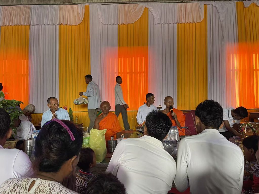 Buddhist monks chanting prayers during Pchum Ben Day ceremony in Cambodia, surrounded by villagers offering food and donations inside a decorated hall.